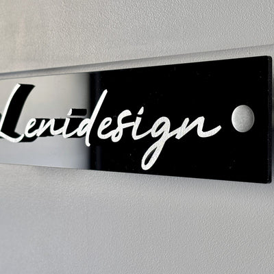 Plaque de porte Leniva posée au mur : plexiglas noir, lettres blanches, entretoises